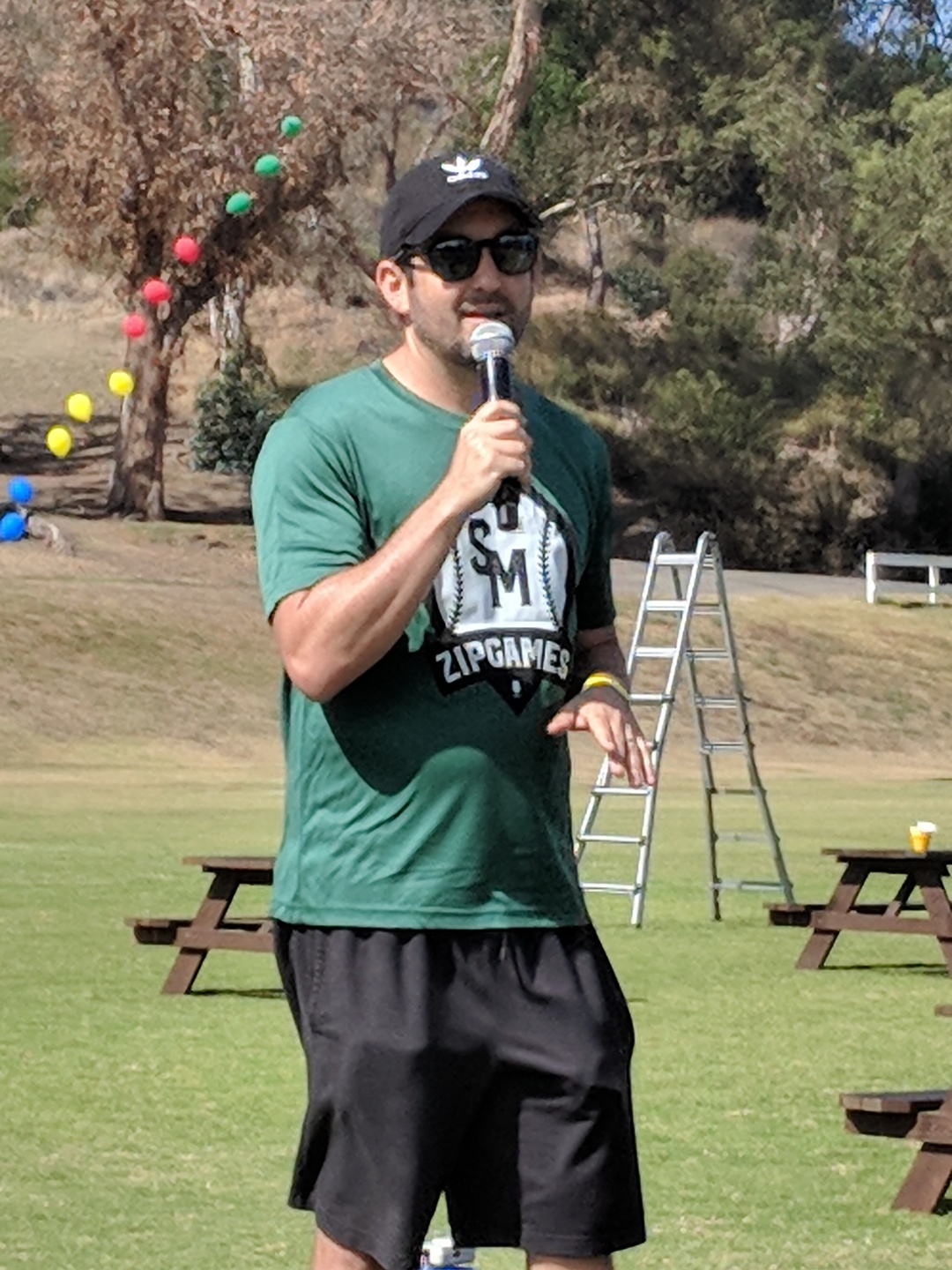 Man in a green shirt and ballcap speaking into a microphone in front of a green field outdoors.