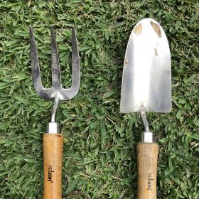 A hand fork and a hand trowel, both with wooden handles, lying side by side on green grass.