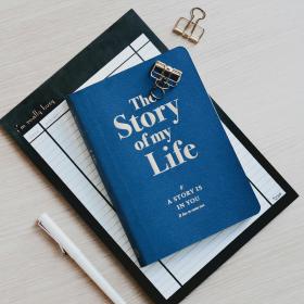 A blue hardcover book titled 'The Story of my Life' resting on a ruled notebook on a pale wood table, with a white pen beside it